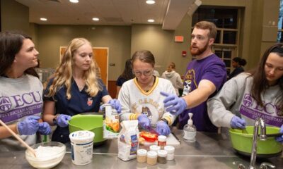 A bearded man in a purple T-shirt talks to a group of women preparing food in a large meeting room.